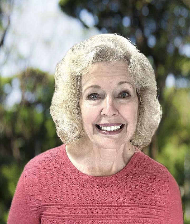 Camille Cornaby smiling outdoors in a coral top, with green trees behind her